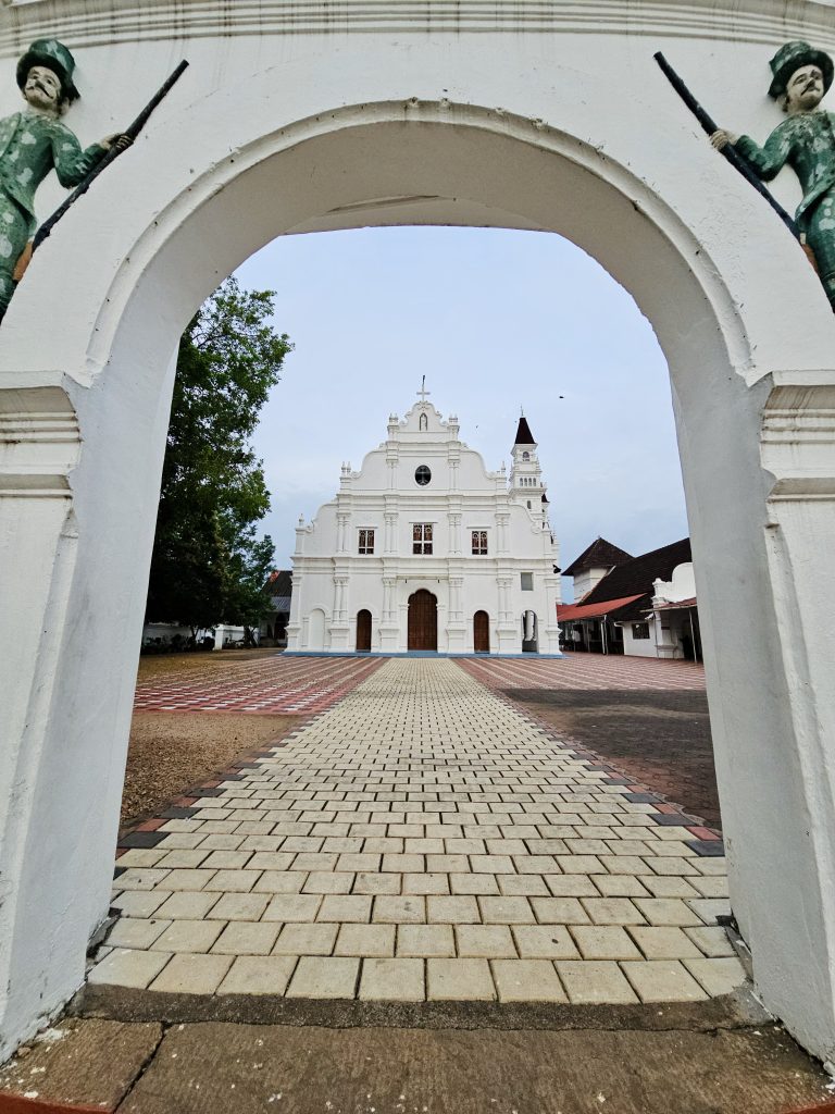 A white historic church viewed through a large arched gateway, with two sculpted figures framing the entrance. The paved pathway leads toward the ornate church façade. Location: Ramapuram, Kottayam, Kerala.
