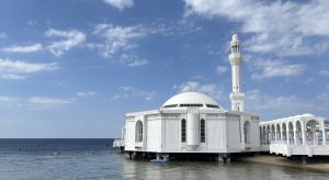 White Al-Rahma floating mosque in Jeddah, Saudi Arabia, standing on pillars above the calm Red Sea, with a large dome, a tall slender minaret and an arched white walkway under a blue sky with soft clouds.
