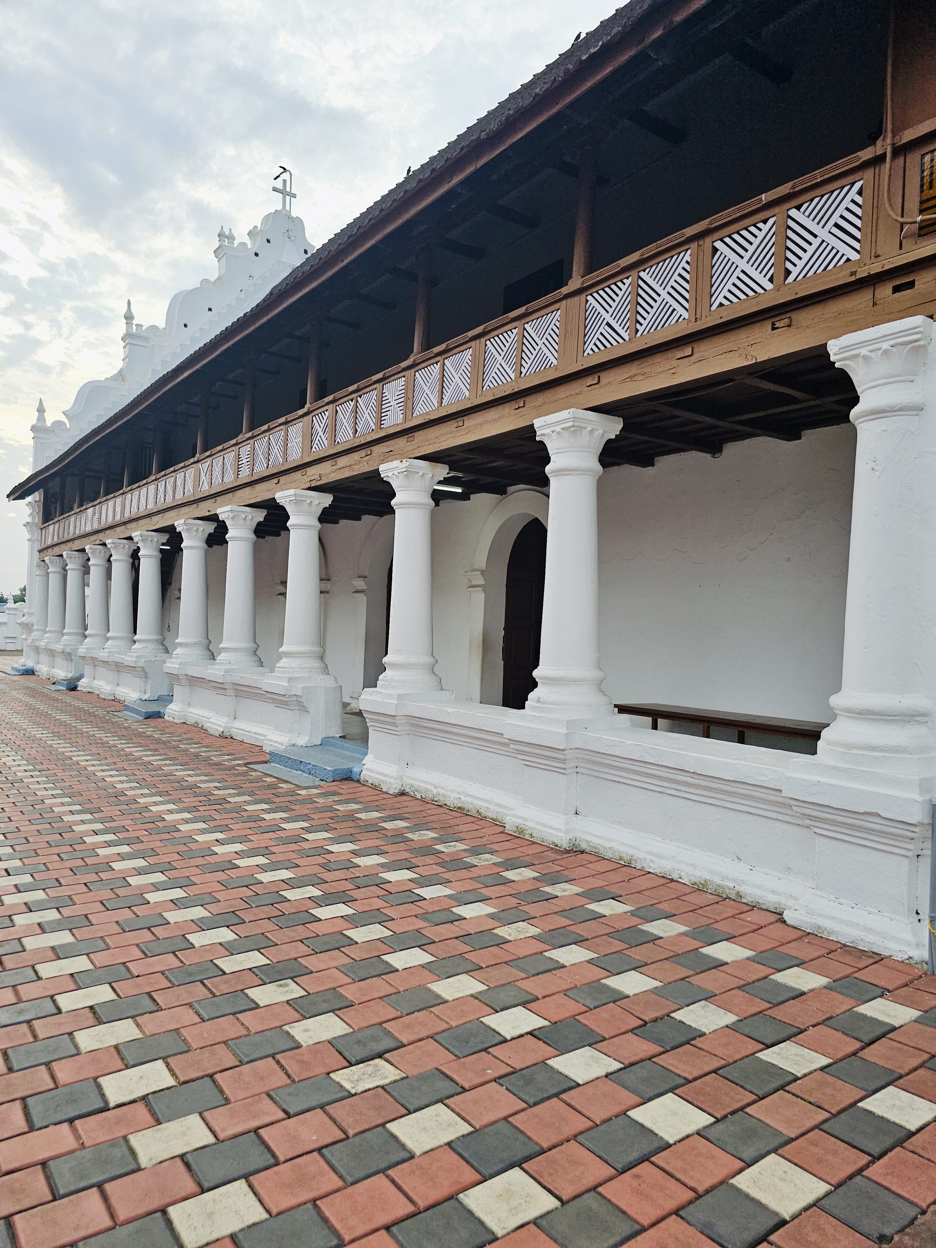 A long white colonnade with traditional wooden railings on the upper level, viewed at an angle. The repeating columns and patterned paving create strong leading lines. Location: Ramapuram, Kottayam, Kerala.