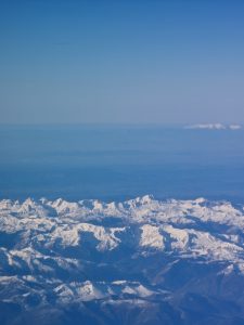 High-altitude aerial photograph showing a vast range of sharp, snow-covered mountain peaks stretching across the horizon under a clear blue sky.
