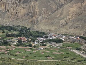 A mountain village with traditional white and red buildings nestled at the base of dramatic, eroded canyon cliffs, surrounded by green fields and patchy vegetation.