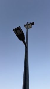 Tall dark metal streetlamp with two rectangular LED light fixtures pointing in different directions against a clear blue sky.
