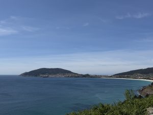 Panoramic view of Cape Finisterre and the Atlantic coast, with green vegetation, a coastal town, and a sandy beach under a clear blue sky.