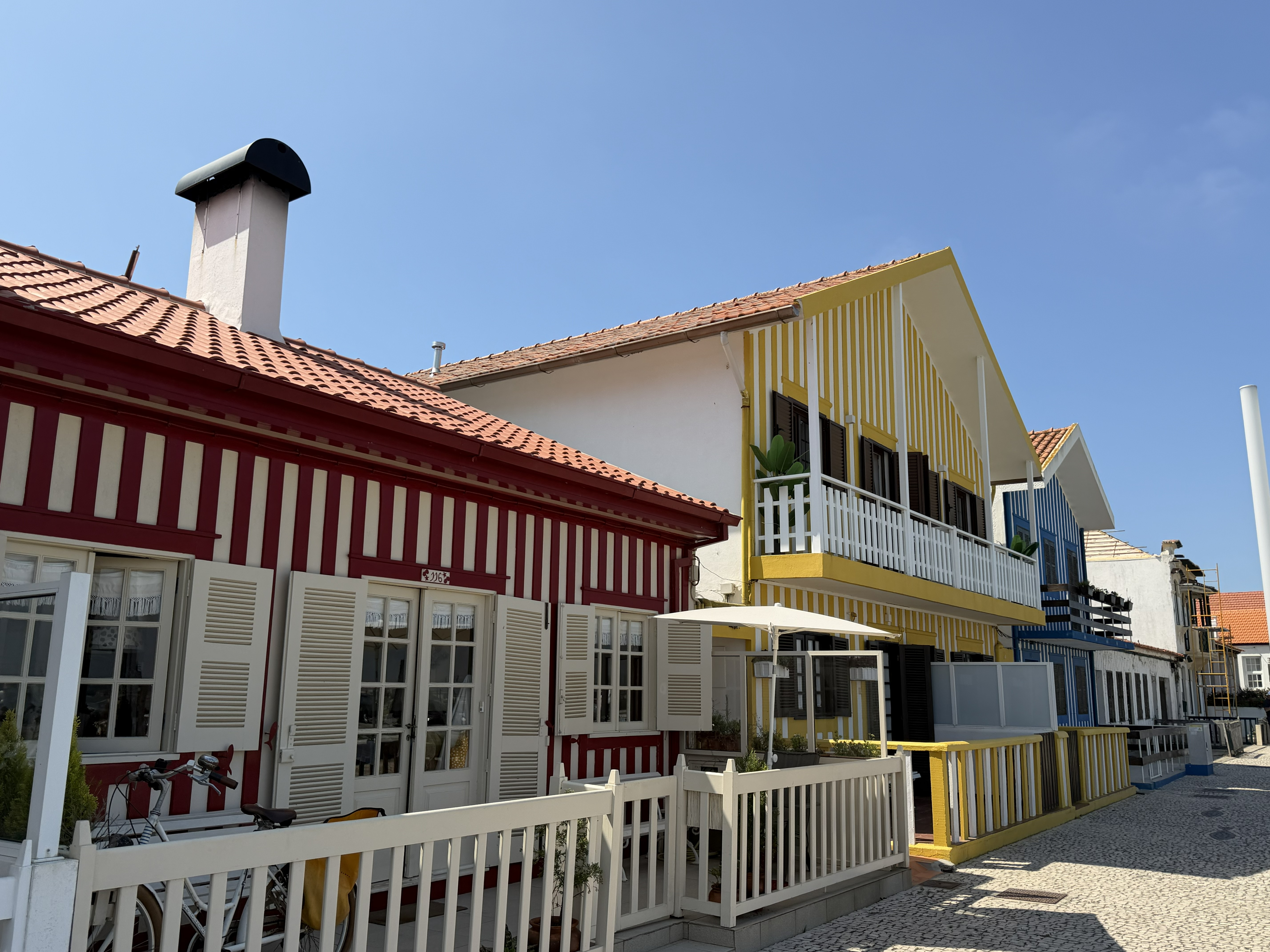 View of the colourful houses of Costa Nova, Portugal, featuring red and white, yellow and white, and blue and white stripes beneath a clear blue sky.