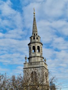 Tall stone church spire reaching into a cloudy blue sky framed by bare tree branches at the bottom.