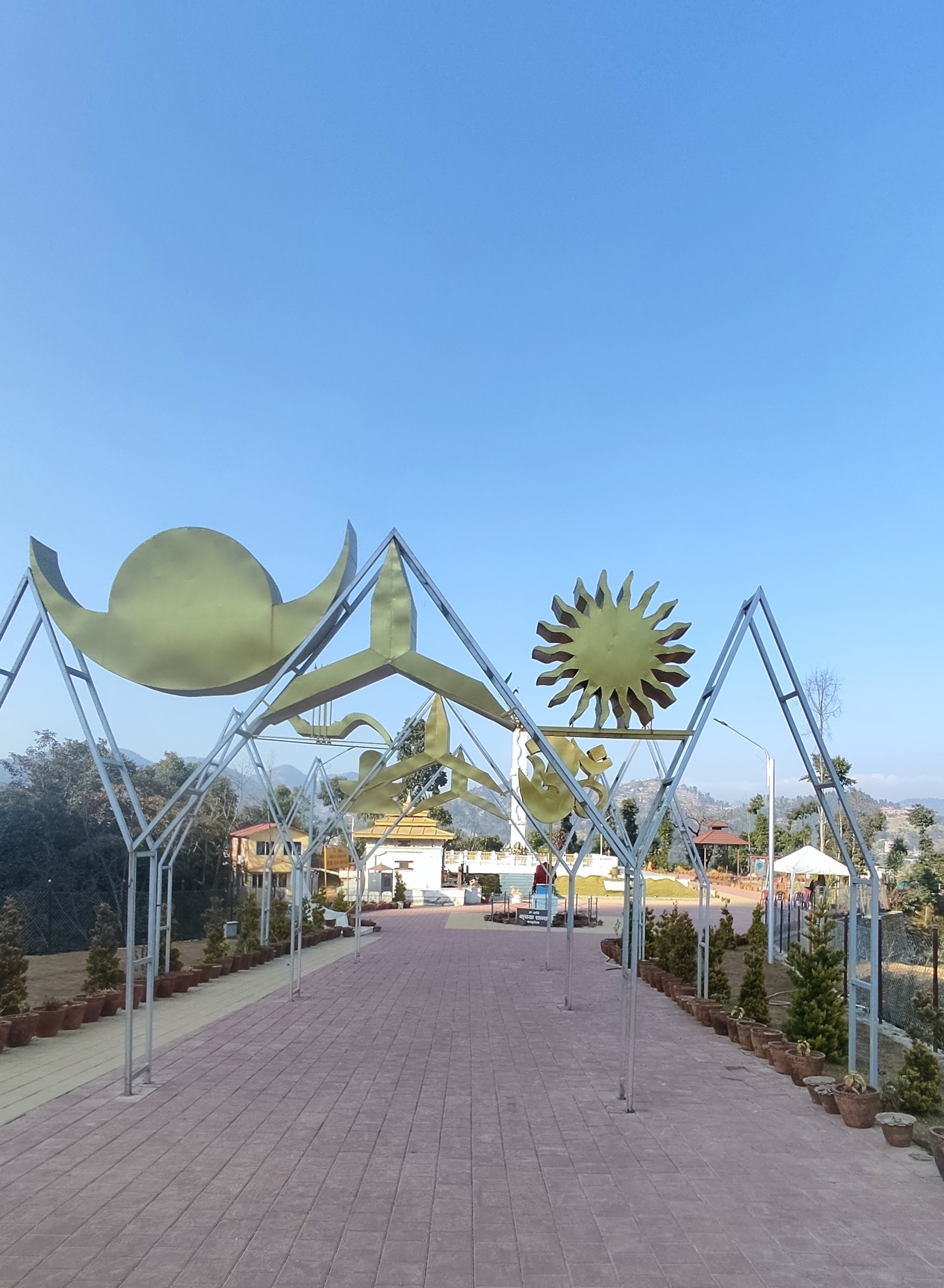 Wide pathway with potted plants and celestial sculptures at Aaditya Mokshya Dham, with green hills and a blue sky in the background.