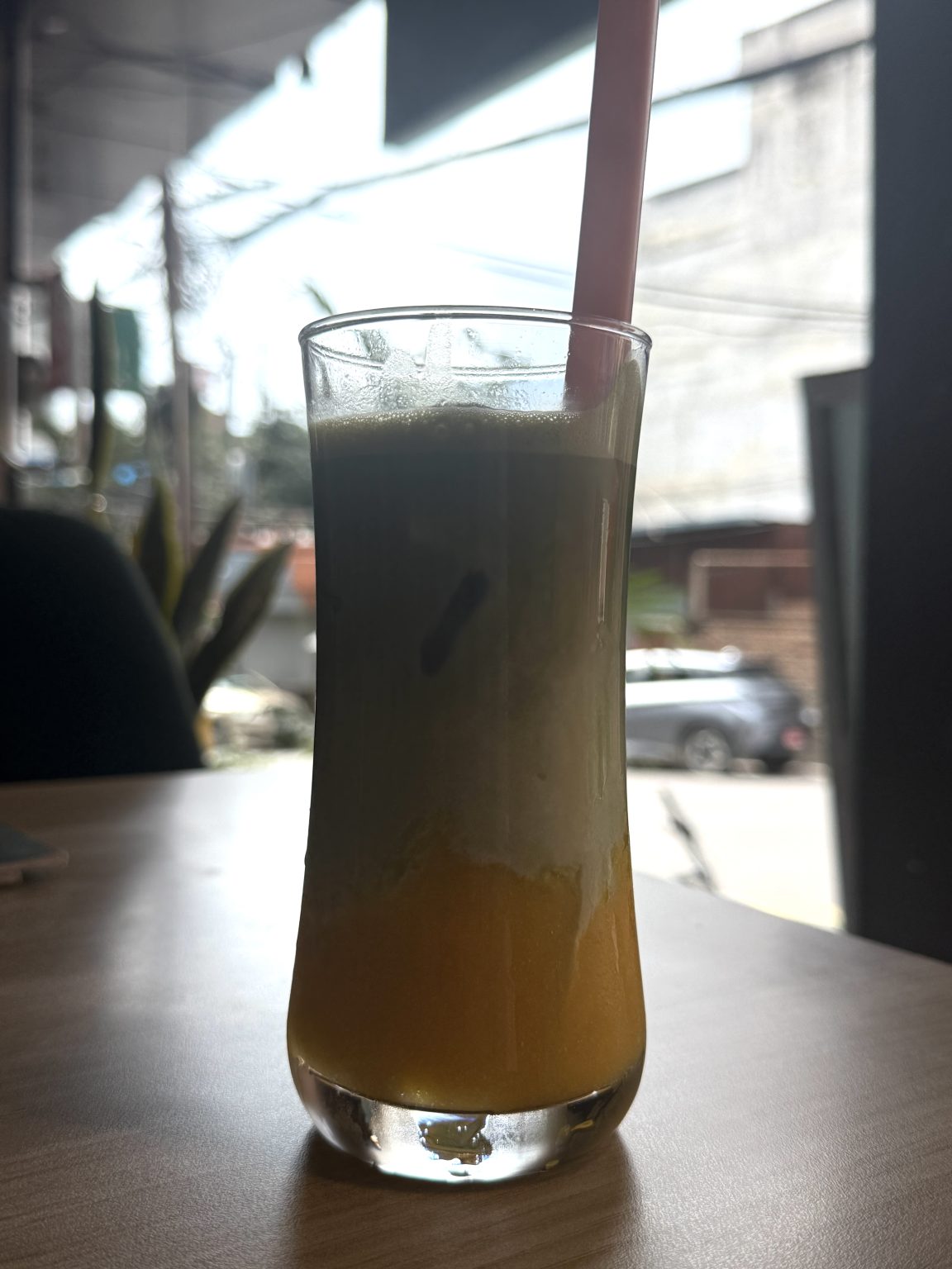 Layered matcha drink with orange, creamy white, and green layers in a clear glass with a pink straw on a wooden table.