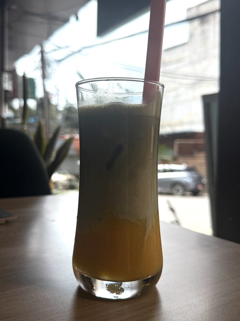 Layered matcha drink with orange, creamy white, and green layers in a clear glass with a pink straw on a wooden table.