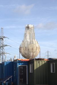 Outdoor public art installation featuring a large wire mesh lightbulb sculpture mounted on top of blue and green shipping containers.