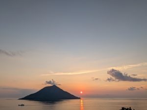 A small island, Hachijo-kojima, silhouetted against an orange sunset sky, with the sun reflecting on the calm sea and a ferry passing on the left.