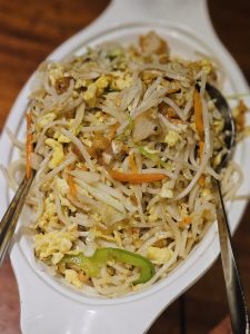 A close-up view of a freshly prepared noodle dish with vegetables, scrambled egg, and pieces of chicken, served on a white plate. The shallow depth of field highlights the food and its textures. Location: Edavannappara, Malappuram, Kerala.