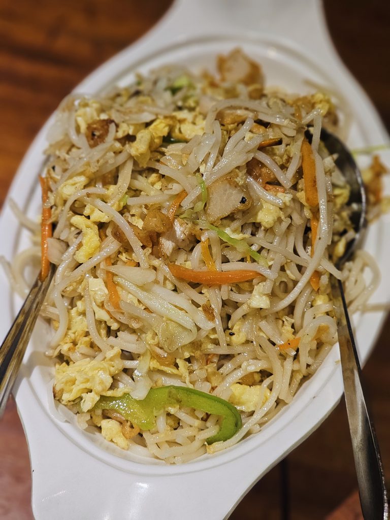A close-up view of a freshly prepared noodle dish with vegetables, scrambled egg, and pieces of chicken, served on a white plate. The shallow depth of field highlights the food and its textures. Location: Edavannappara, Malappuram, Kerala.