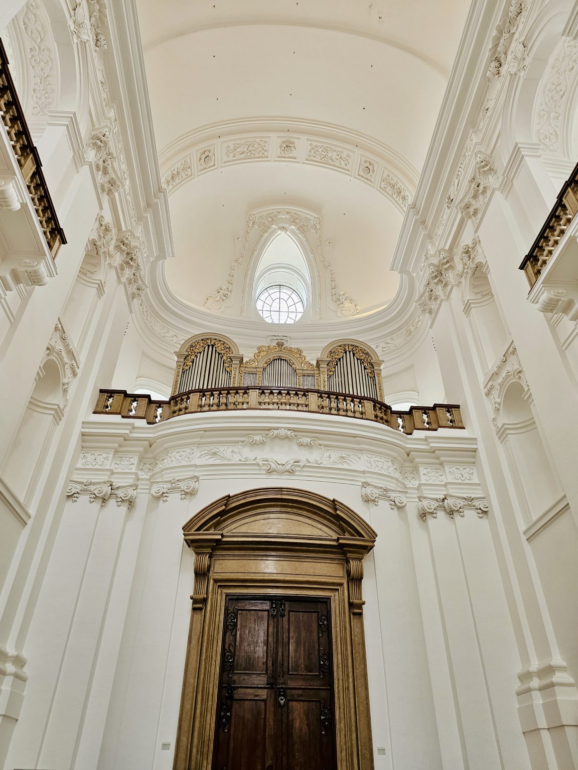 A grand Baroque church interior features an ornate pipe organ, elaborate white stucco decoration, a large arched ceiling, and a historic wooden doorway. Location: Salzburg, Austria.