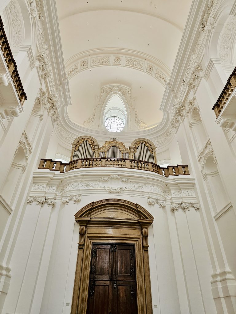 A grand Baroque church interior features an ornate pipe organ, elaborate white stucco decoration, a large arched ceiling, and a historic wooden doorway. Location: Salzburg, Austria.