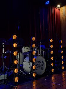 White acoustic drum set on a wooden stage with “Tutto Passa” on the bass drum, warm amber lights, and a dark red curtain in the background.