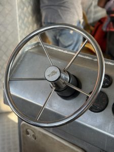 Stainless steel ship’s wheel with six spokes mounted on a metal console with gauges, used for steering a boat. 
