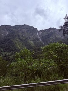 A tall, slender waterfall cascading down a steep, lush green mountain slope framed by dense foliage in the foreground under an overcast sky.