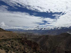 A high-elevation vista showing dramatic, eroded canyon cliffs and sweeping arid valleys under a blue sky scattered with white clouds.