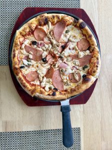 Pizza with a golden-brown crust, melted cheese, pepperoni, chicken, black olives, and green herbs on a dark wooden serving board.