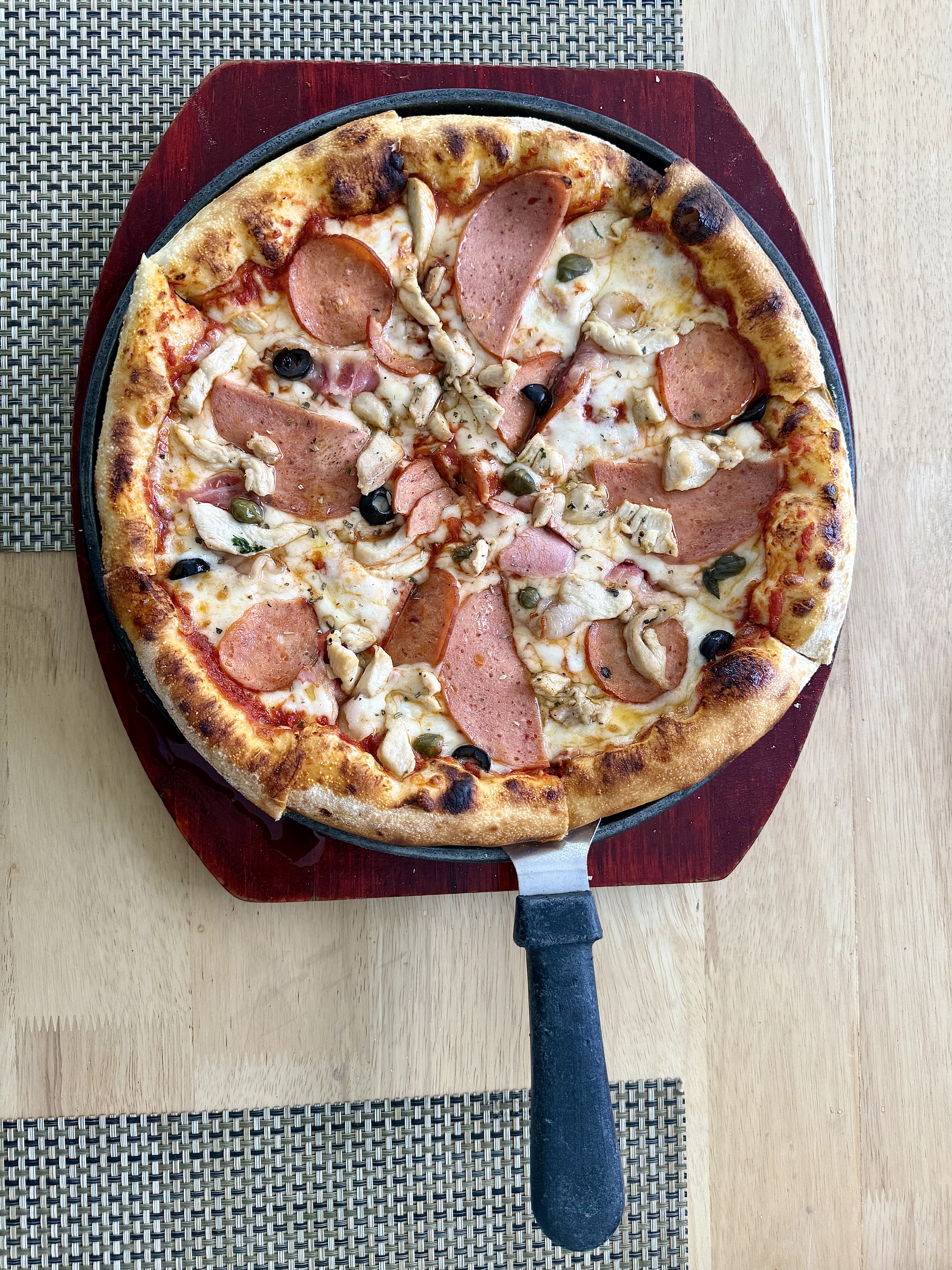 Pizza with a golden-brown crust, melted cheese, pepperoni, chicken, black olives, and green herbs on a dark wooden serving board.