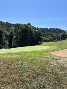 Lush green golf course with a putting green and flag, surrounded by trees and rolling hills under a blue sky.