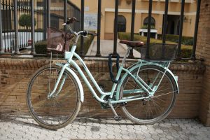 A vintage-style bicycle leans against a brick wall. The bike is painted in a soft mint green color, contrasting with the warm tones of the wall. It features large, thin wheels that show some wear, and there are two baskets: one made of metal mesh in the front, ideal for carrying small items, and a larger one in the back.
