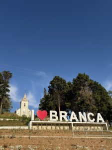 Low-angle view of an “I ❤️ BRANCA” sign on a hill, with a small white church and trees under a clear blue sky.