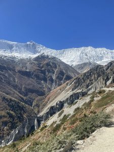 A panoramic vista of rugged, carved canyon walls and steep mountain slopes rising up to a massive, heavily glaciated ridge under a vibrant, cloudless blue sky.