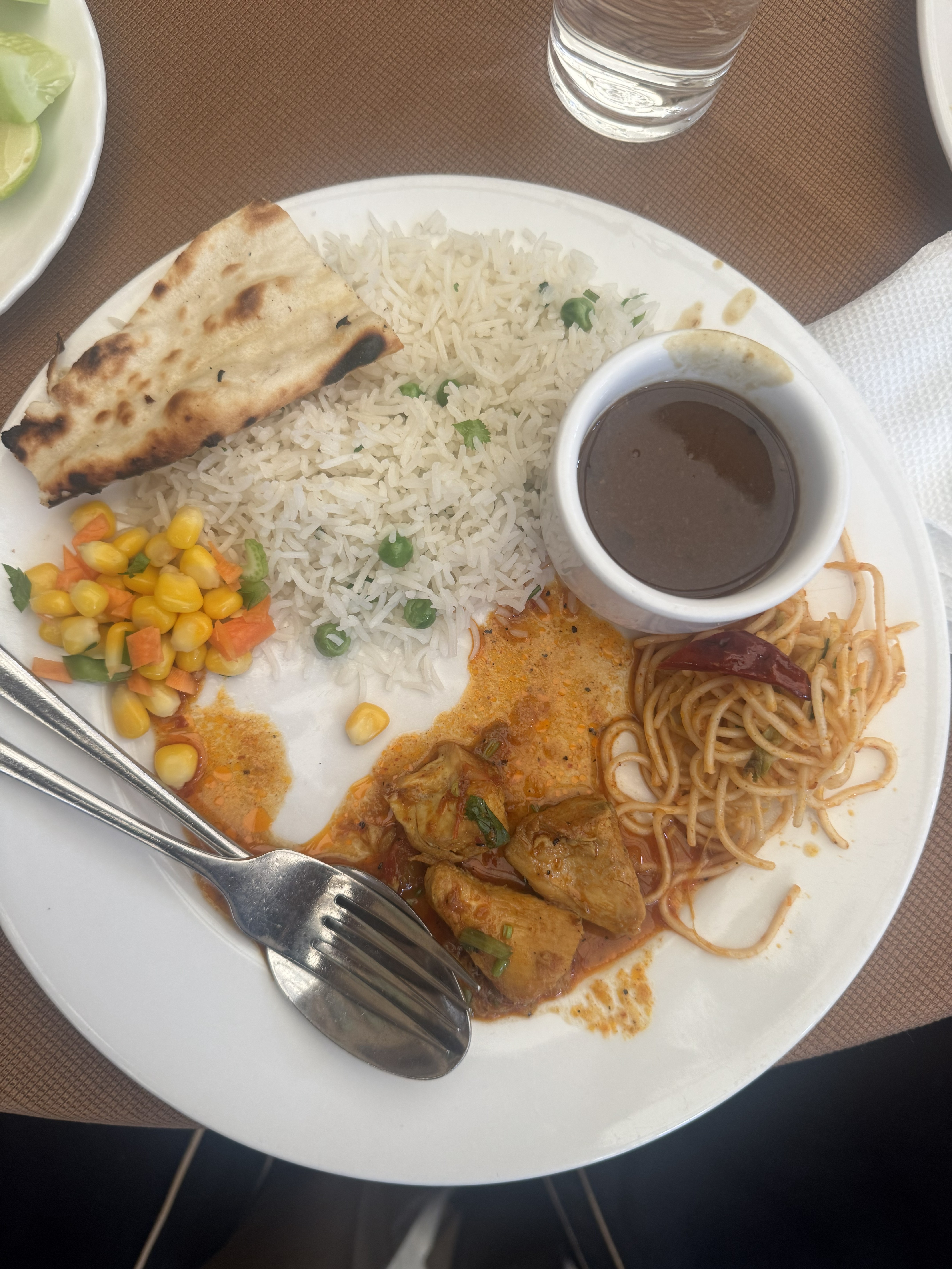 A meal served on a white plate, featuring basmati rice, naand bread, chicken curry, stir-fried noodles, a small corn salad, and a bowl of dark dipping sauce alongside a glass of water.