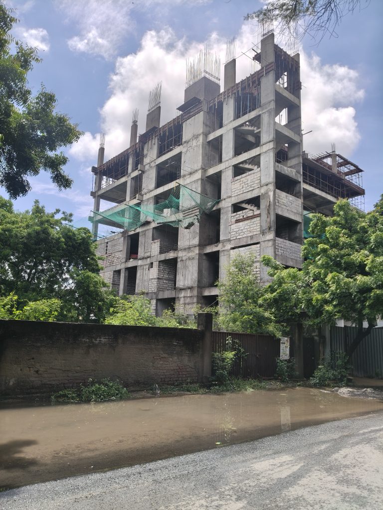 Partially constructed concrete building with scaffolding and green safety netting, surrounded by trees and a gravel road.