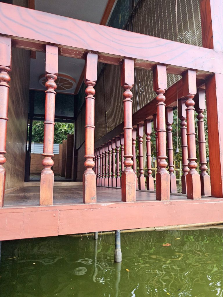 A traditional wooden veranda with rows of turned, red-painted pillars overlooking a body of water. The repeating pillars, reflections, and perspective create an attractive architectural composition. Location: Ayamkudy, Kottayam, Kerala.