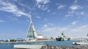 Tall glass-and-white sail-shaped tower of Jeddah Yacht Club beside its calm marina, with a large black-and-white ship moored on the right, a rocky breakwater in front and a wide blue sky with wispy clouds.
