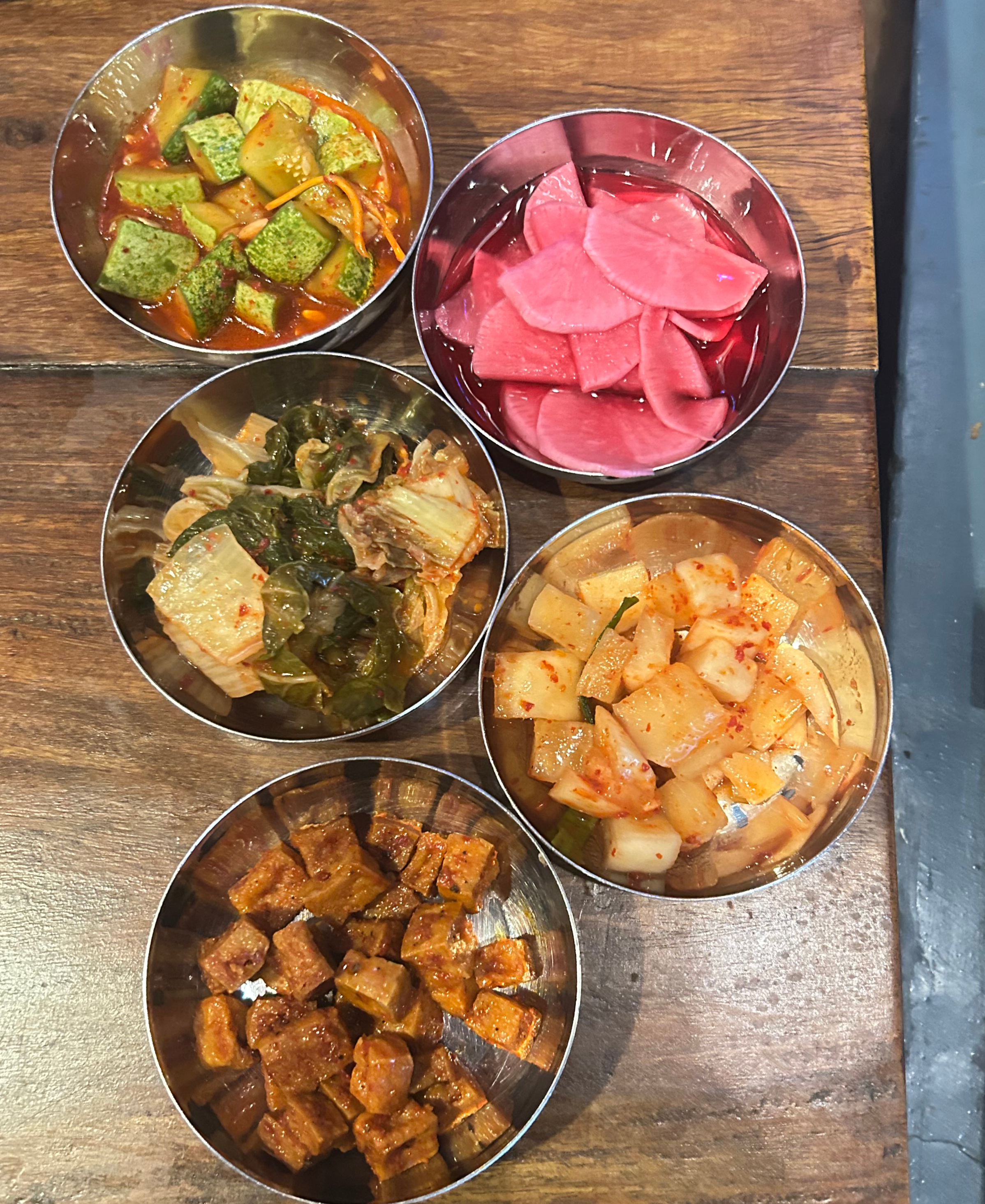 Five small metal bowls filled with colorful vegetable side dishes and sauces on a wooden table.