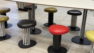 Modern stools with rounded triangular seats in red, black, and olive green, resting on solid black or black-and-white striped bases beneath a white table.
