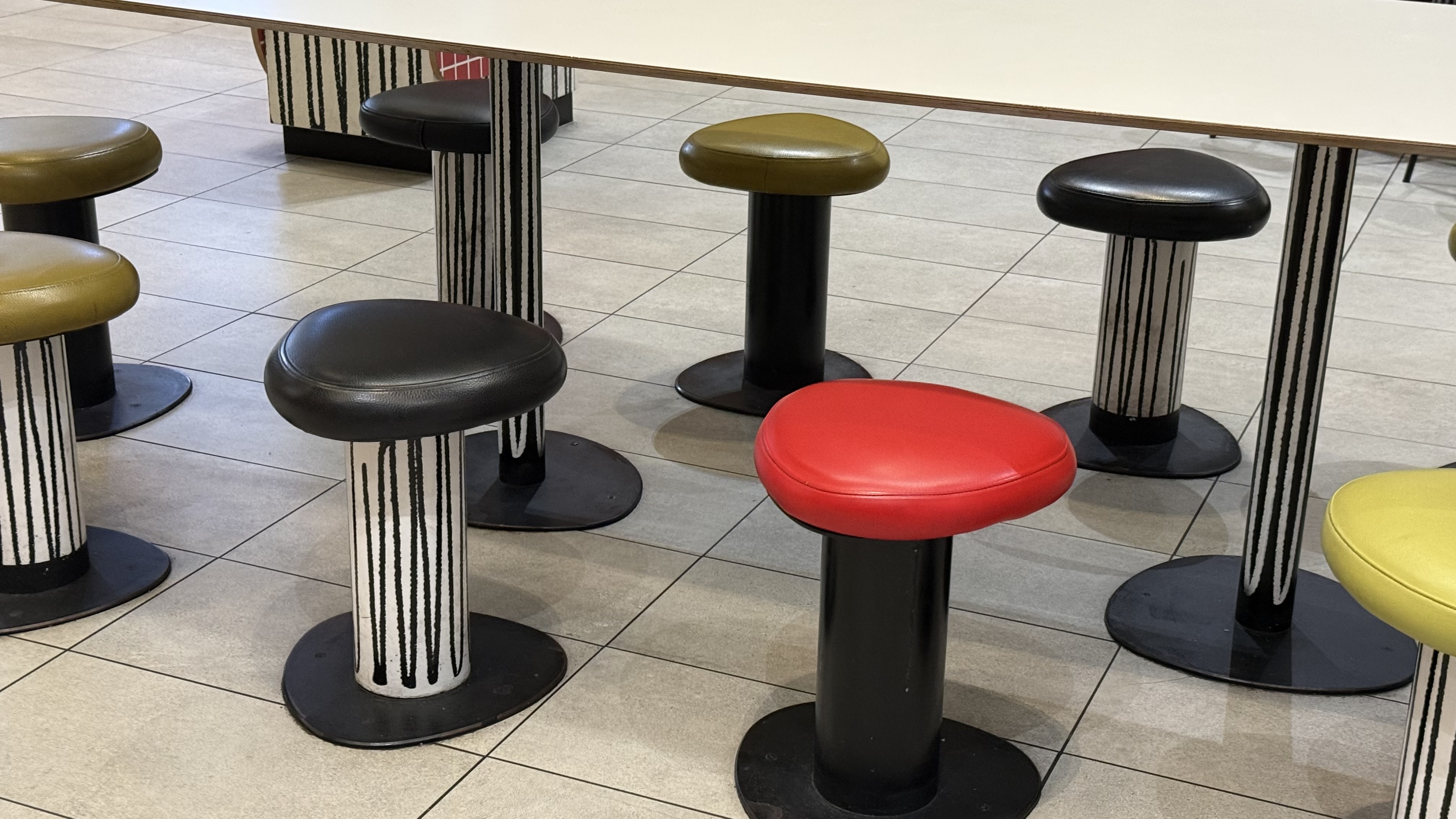 Modern stools with rounded triangular seats in red, black, and olive green, resting on solid black or black-and-white striped bases beneath a white table.