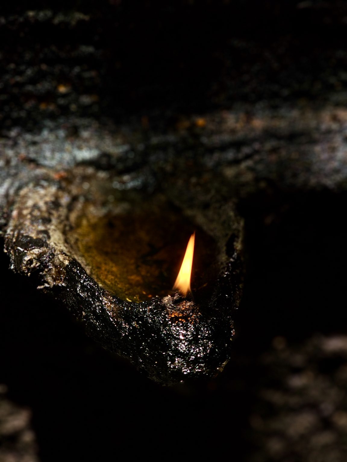 A single oil-lamp flame glows from a traditional lamp in a dark, natural setting. Low-key lighting and shallow focus create a quiet, intimate, atmospheric mood. Location: Ayamkudy, Kottayam, Kerala.