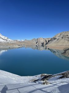 A high-altitude Alpine lake (Tilicho Lake) with deep, calm turquoise-blue water reflecting surrounding snow-dusted, arid mountain slopes beneath a bright blue sky.