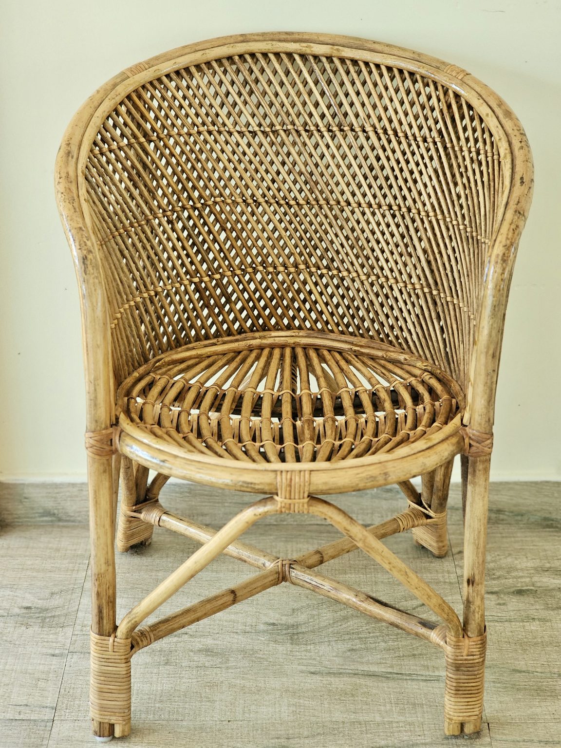 A well-composed front view of a traditional cane chair, showcasing its intricate woven back, curved frame, crossed supports, and craftsmanship. The soft natural tones complement the rustic character of the chair. Location: Ayamkudy, Kottayam, Kerala.