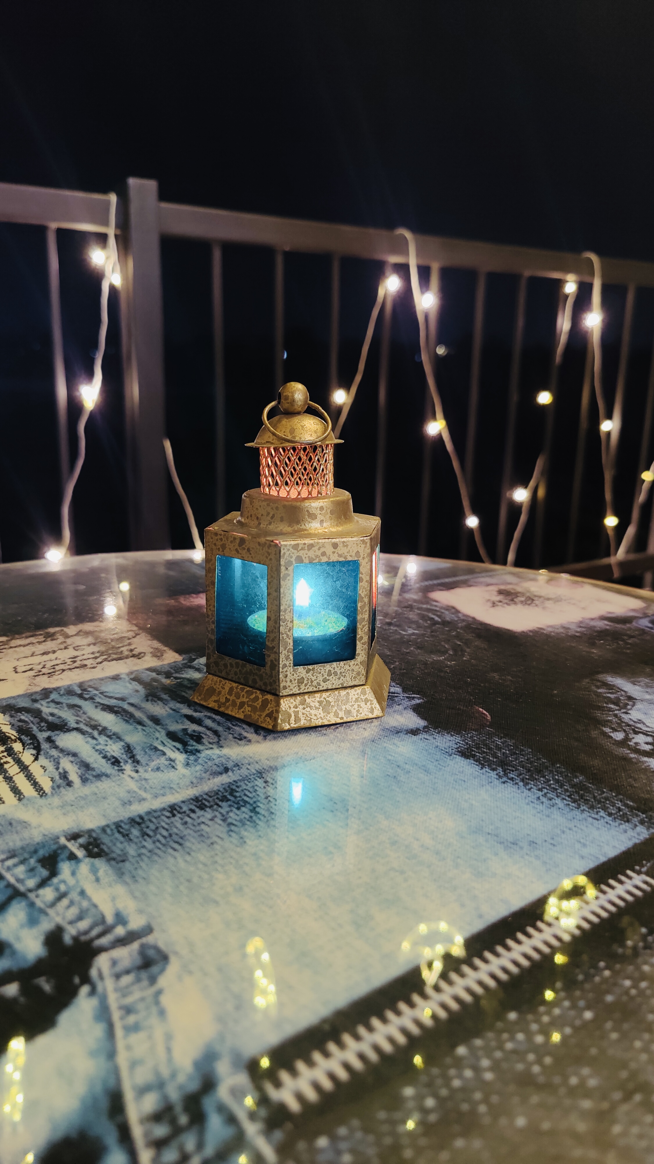 A decorative lantern glowing blue sits on a table at night, surrounded by warm white string lights along a balcony railing.
