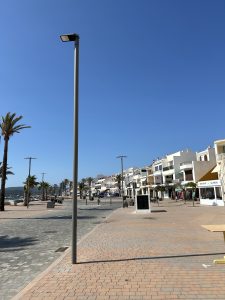 A wide paved seaside promenade with streetlights, palm trees, and white buildings under a clear sky.