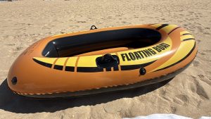 Orange and yellow inflatable boat with a black interior, resting on the dry sand of a beach. The boat has the text 'FLOATING BOAT' printed on its side and features black oar mounts.
