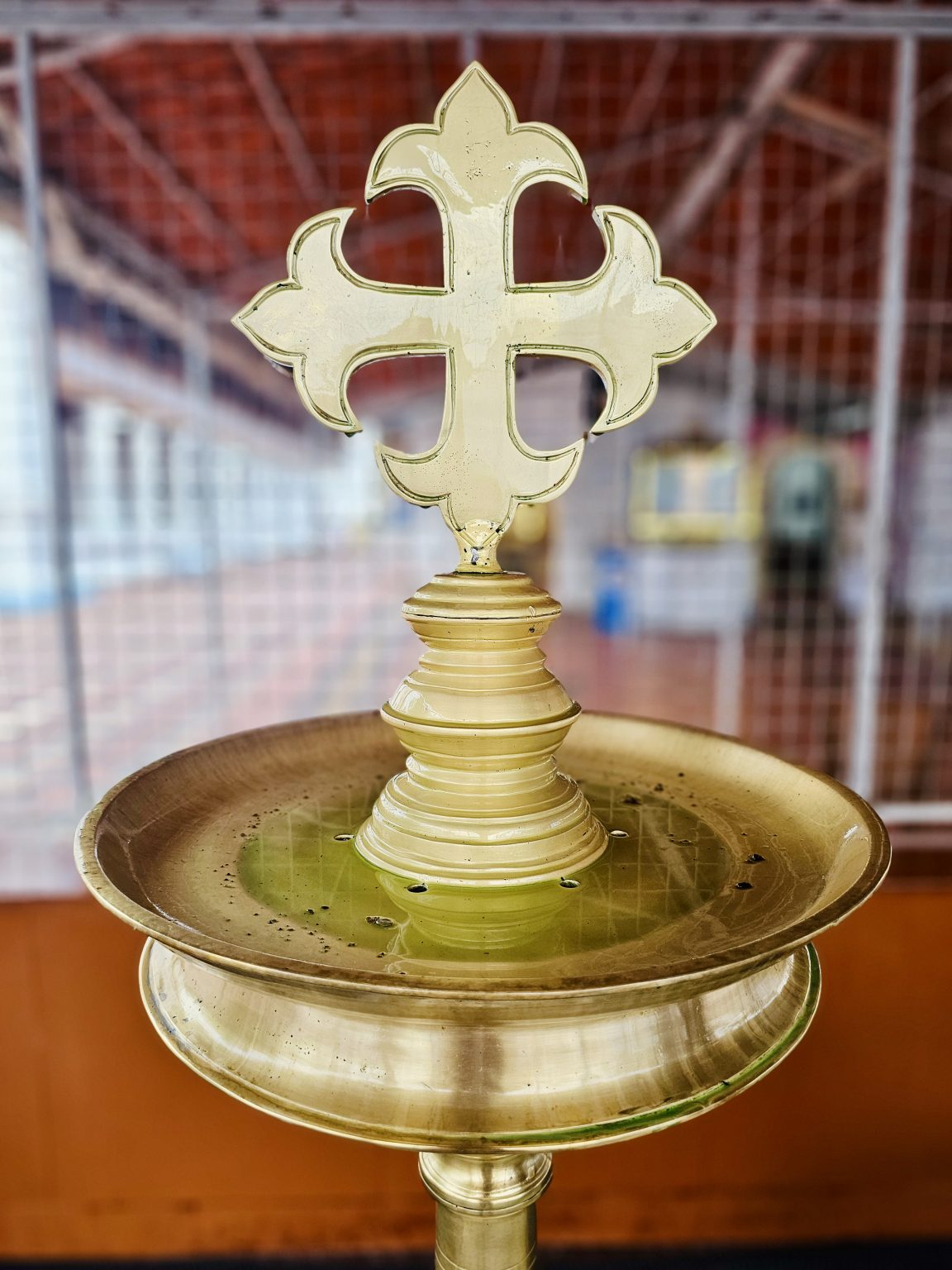 An ornate golden cross and brass vessel captured against a softly blurred traditional interior. The detailed craftsmanship and reflective metal surface create a strong subject. Location: Ramapuram, Kottayam, Kerala