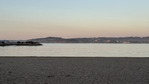 View of a calm sea from a sandy beach at dusk, showing a distant coastline with hills and a stone breakwater.
