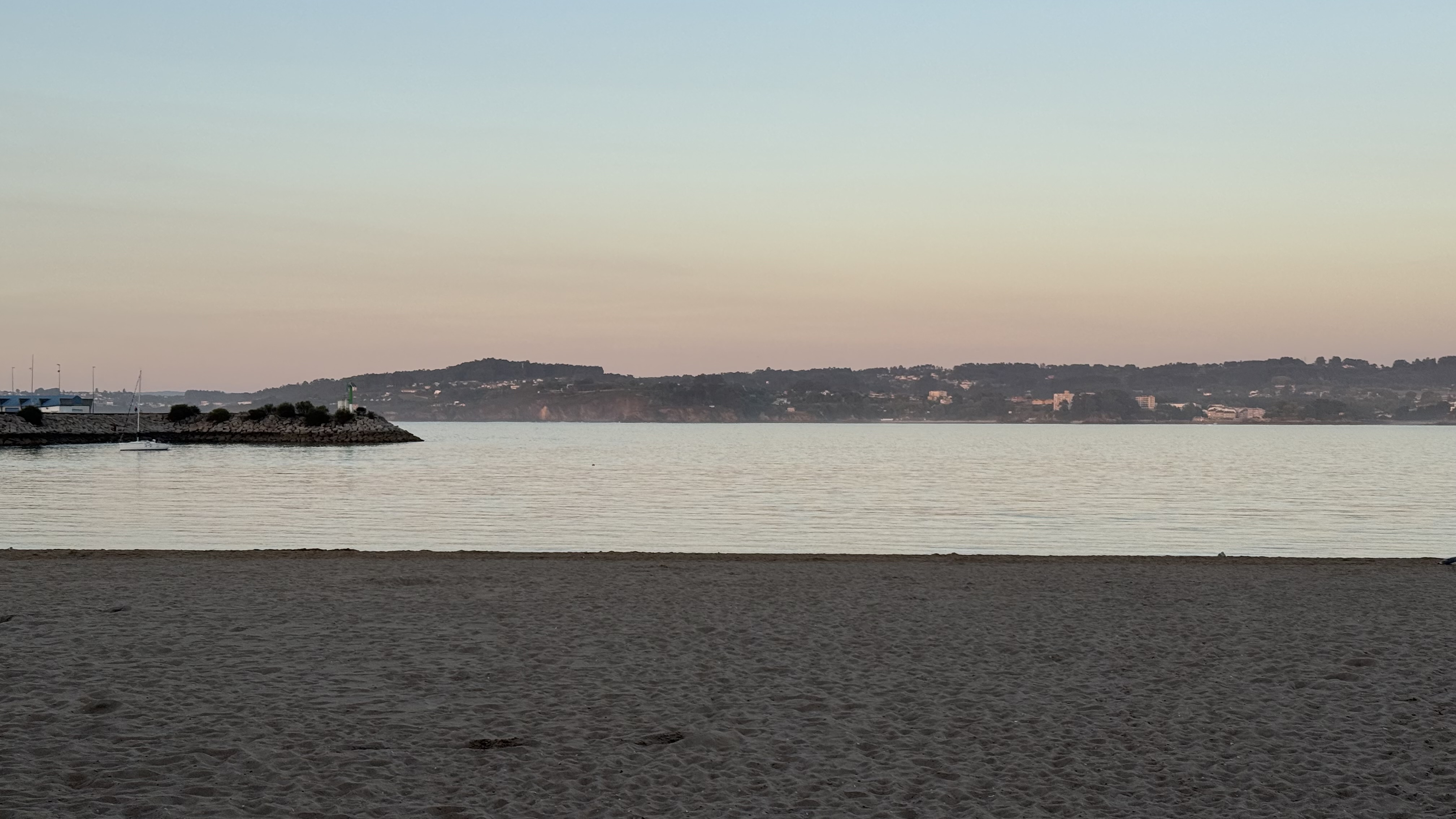 View of a calm sea from a sandy beach at dusk, showing a distant coastline with hills and a stone breakwater.