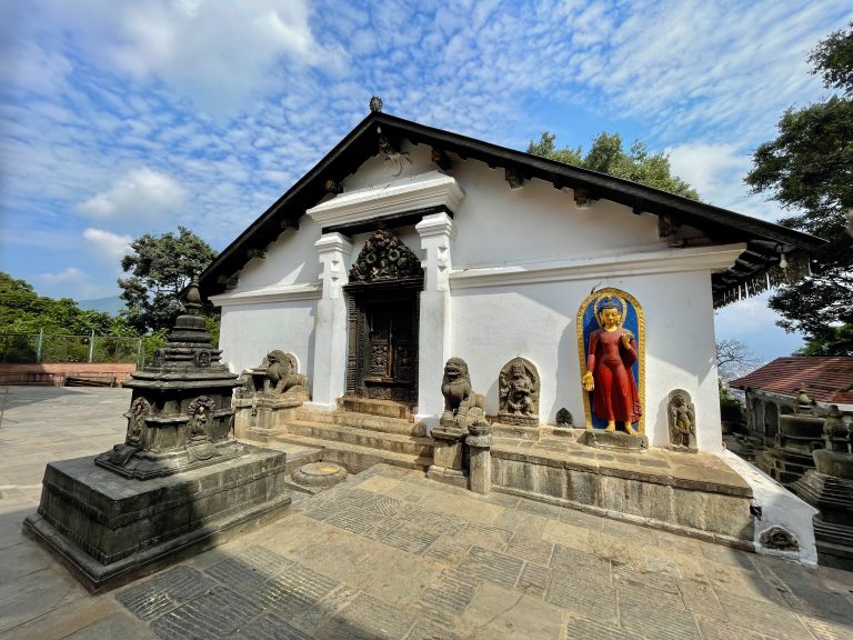 A traditional Buddhist temple (Shantipur Shrine) with white walls, intricate wooden carvings, stone sculptures, and a colorful Buddha figure stands in a historic courtyard. A blue sky and surrounding trees add to the peaceful setting. Location: Swayambhunath complex, Kathmandu, Nepal.