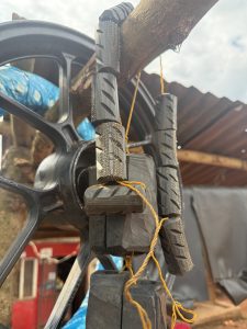 Blocks of old motorcycle tire treads tied with yellow rope hang from a wooden beam in an outdoor workshop, with a metal wheel and a small tin-roof structure in the background. #WCJinja
