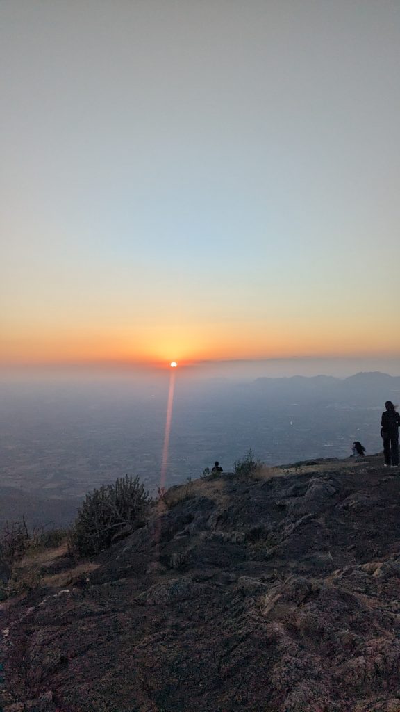 Rocky mountain viewpoint at sunset, with two people silhouetted against distant mountain ranges and an orange and yellow sky.