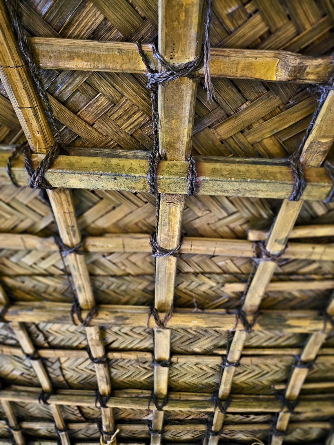 A detailed view of a traditional woven palm-leaf roof supported by bamboo poles tied together with natural fiber. The repeating geometric patterns and warm, earthy textures showcase traditional construction. Location: Ayamkudy, Kottayam, Kerala.