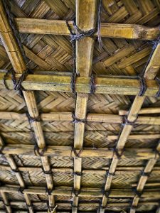 A detailed view of a traditional woven palm-leaf roof supported by bamboo poles tied together with natural fiber. The repeating geometric patterns and warm, earthy textures showcase traditional construction. Location: Ayamkudy, Kottayam, Kerala.