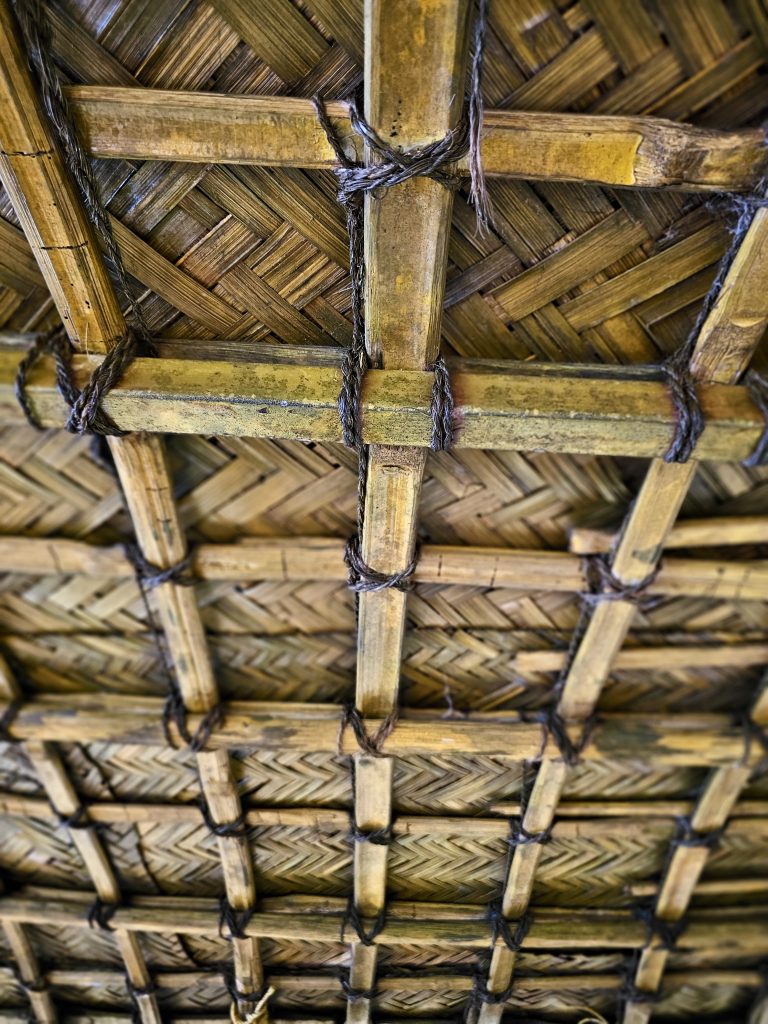 A detailed view of a traditional woven palm-leaf roof supported by bamboo poles tied together with natural fiber. The repeating geometric patterns and warm, earthy textures showcase traditional construction. Location: Ayamkudy, Kottayam, Kerala.